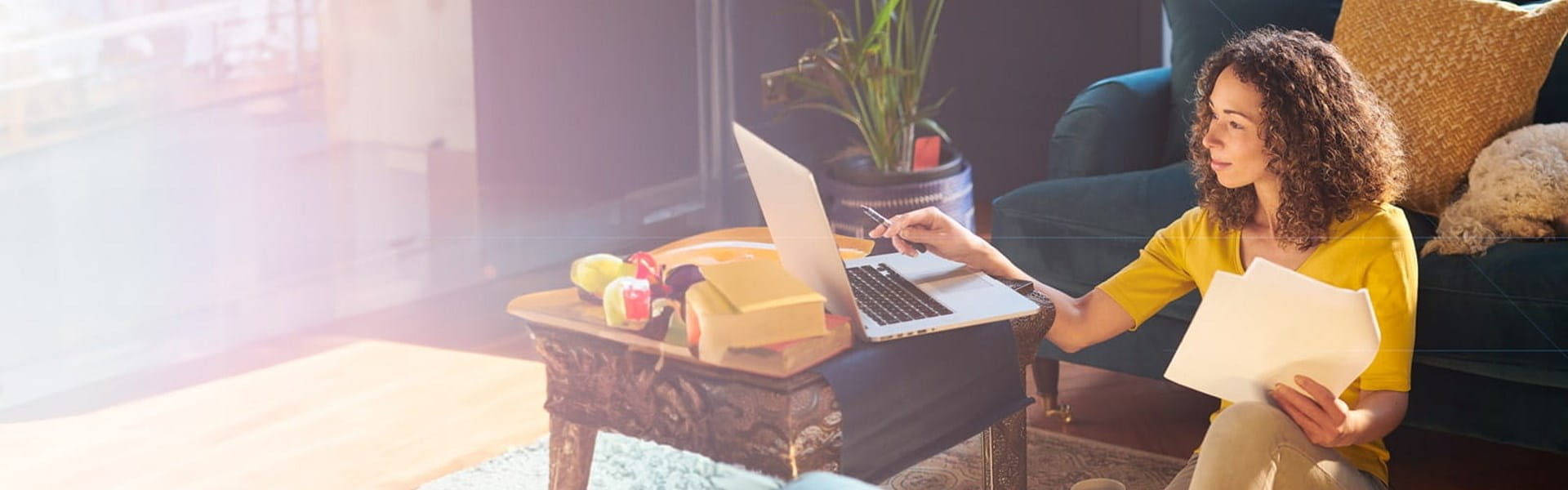 female on laptop working while sitting on the floor and holding papers in her hand