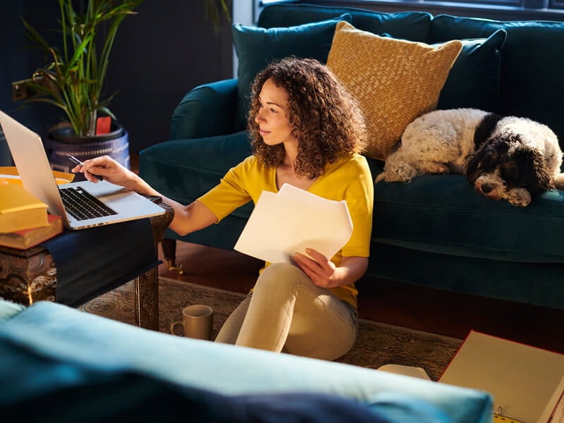female on laptop working while sitting on the floor and her dog is next to her on the couch