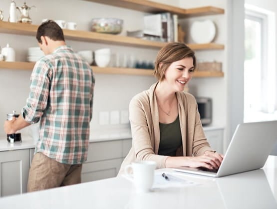 female at a laptop in the kitchen while male spouse prepares something behind her
