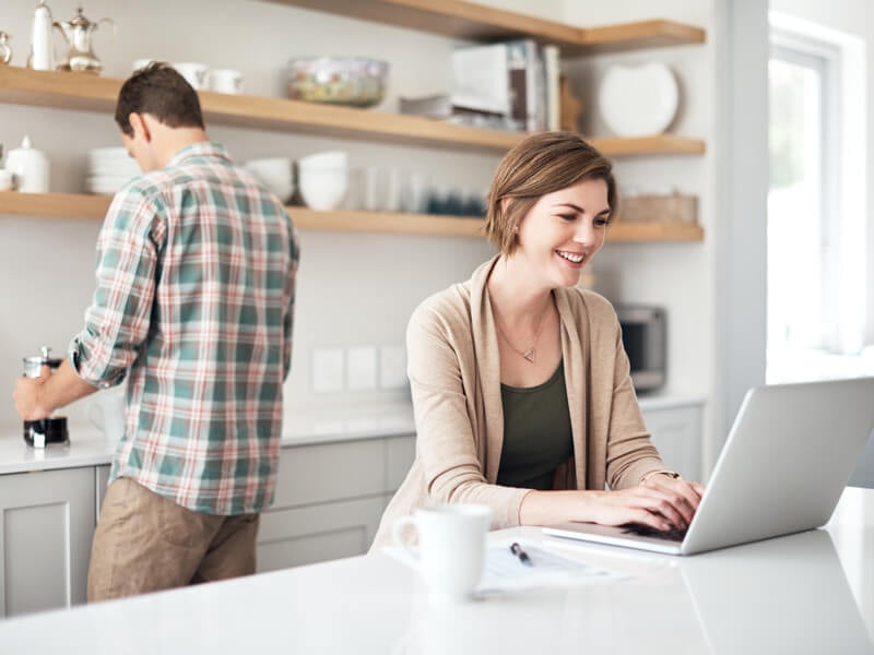 female at a laptop in the kitchen while male spouse prepares something behind her