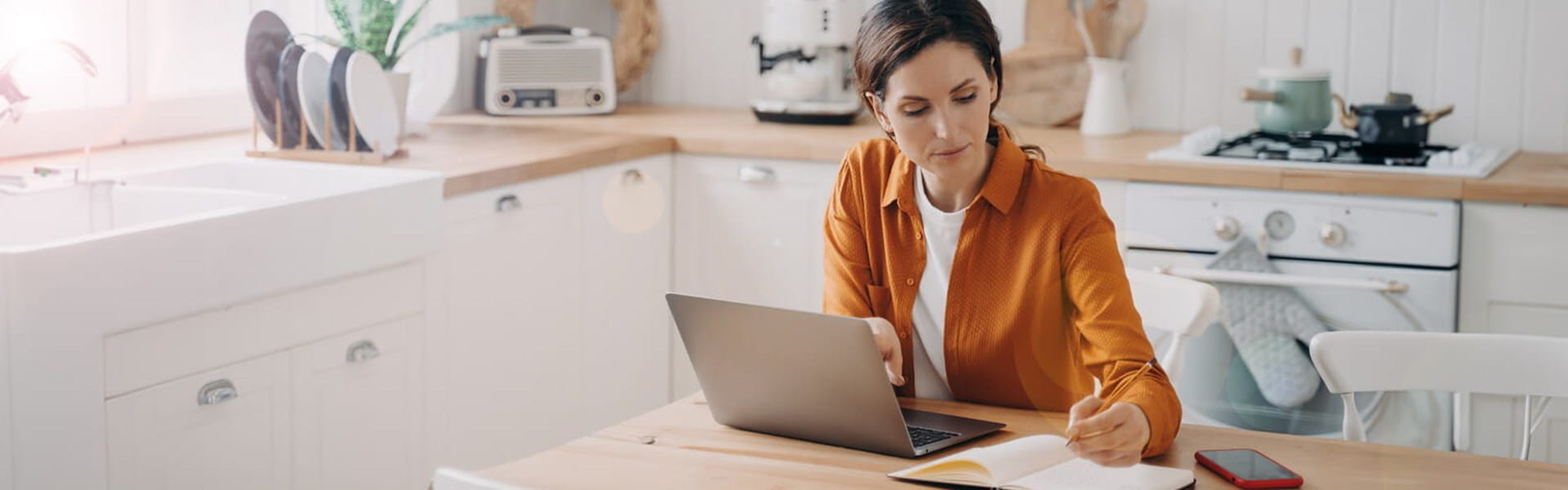 woman in orange shirt on her laptop taking notes in her kitchen