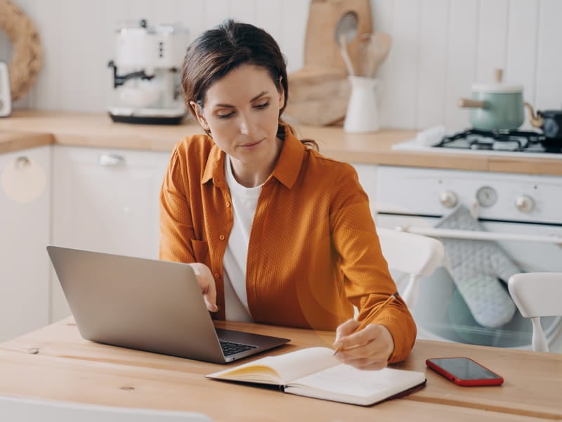 woman in orange shirt on her laptop taking notes in her kitchen