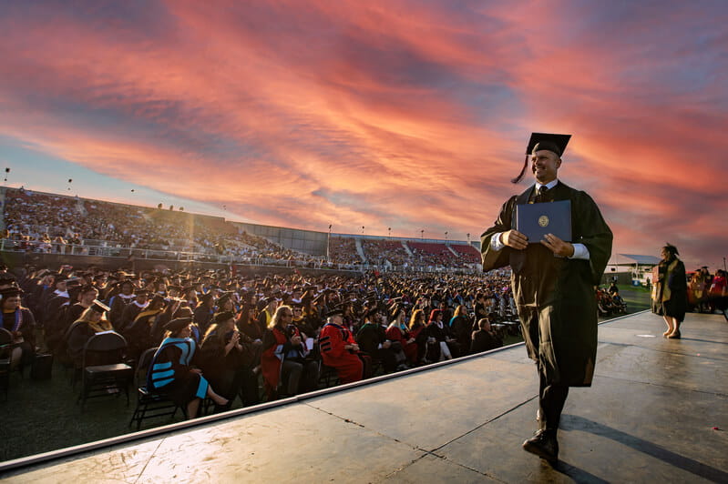 UMass Global Graduate walking across the stage with audience in the background