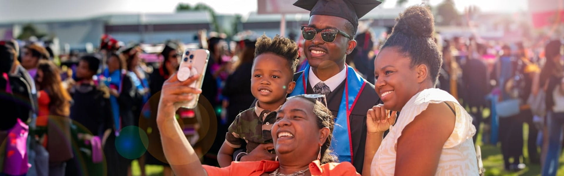 male UMass Global graduate at commencement taking a selfie with his family