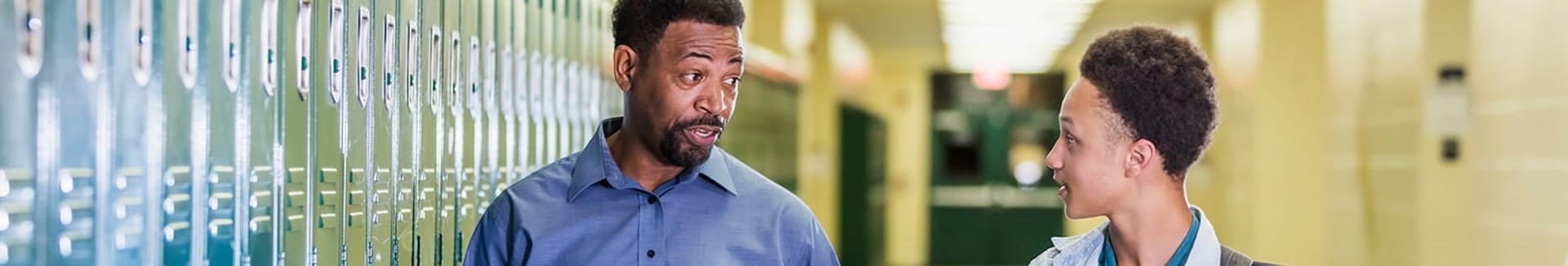 male school counselor supporting  male student walking through school halls by locker