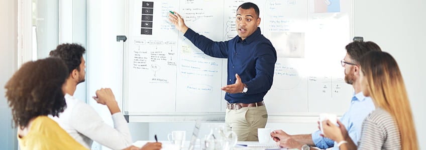 Man standing at white board presenting to a group of colleagues.