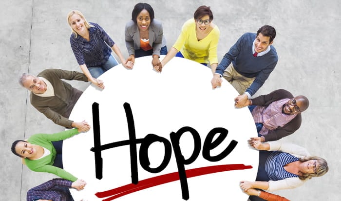 An overhead view of a diverse group of people encircling a round sign emblazoned with the word "Hope," which is designed in a handwriting-style font.