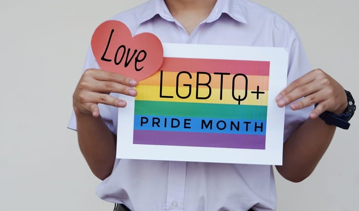 A man holding a paper heart emblazoned with the word "Love" over a sign with the message "LGBTQ+ Pride Month" superimposed over a depiction of a rainbow flag.