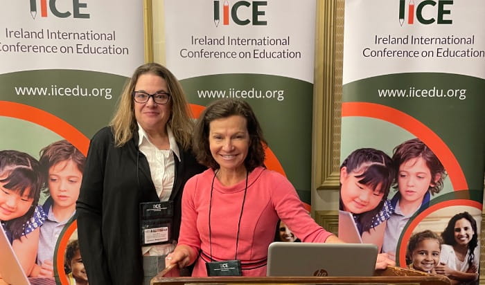 UMass Global faculty members Dr. Kimberly Greene and Dr. Jessica Bogunovich stand side by side behind a podium. Behind them are three identical posters that read "IICE Ireland International Conference on Education" and also contain the URL www.iicedu.org."
