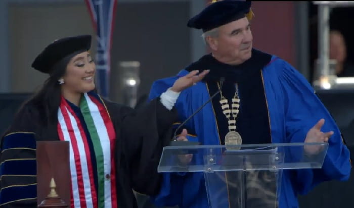 Grad and academic advisor Stephanie Herrera stands next to Chancellor David Andrews on the stage during the 2022 Southern California Commencement. She is pointing toward her parents, who are out of the picture. Both Herrera and Andrews wear graduation regalia.