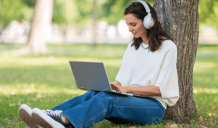 A woman wearing headphones using a laptop while sitting with her back to a tree trunk.