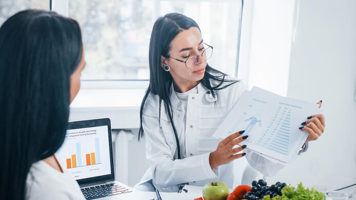 Female nutritionist with laptop gives consultation to patient indoors in the office.