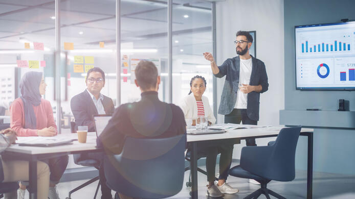 Diverse Modern Office: Businessman Leads Business Meeting with Managers, Talks, uses Presentation TV with Statistics, Infographics. Digital Entrepreneurs Work on e-Commerce Project.