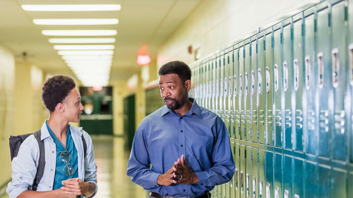 An African-American high school student walking with his teacher or school principal, in the hallway by a row of lockers. They are talking. The teacher has a serious expression on his face, a mature man in his 50s. The male student is an 18 year old teenage boy carrying a backpack.