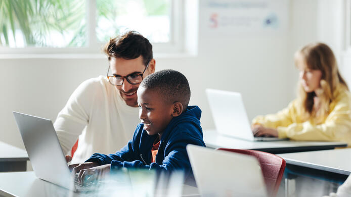 Male teacher helps a young boy with computer-based learning in a classroom setting. Child tutor providing a lesson in an elementary school, with a focus on coding and basic digital literacy.