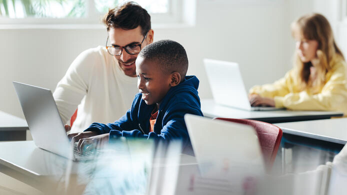 Male teacher helps a young boy with computer-based learning in a classroom setting. Child tutor providing a lesson in an elementary school, with a focus on coding and basic digital literacy.
