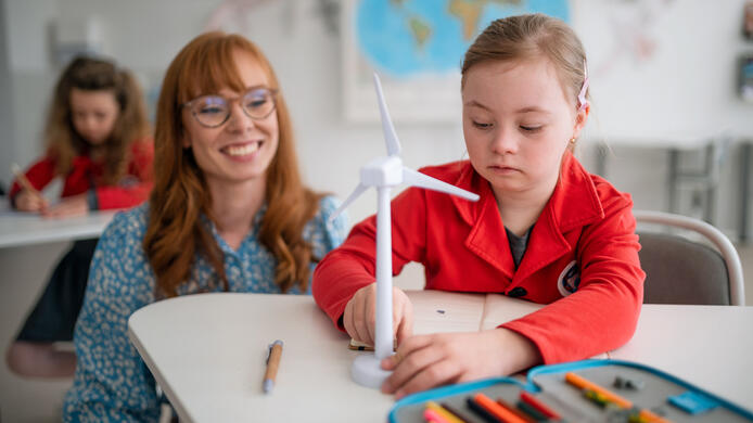 A down syndrome schoolgirl with model of wind turbine with help of teacher learning about eco-friendly renewable sources of energy in class at school, integration concept.