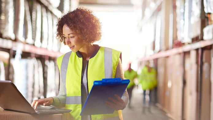 female supply chain manager in a yellow safety vest checking inventory on a computer