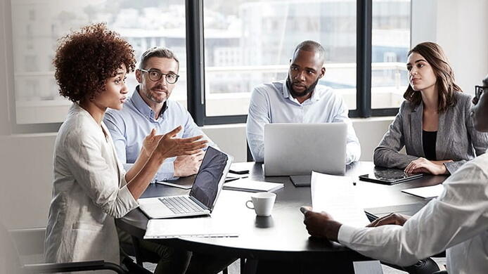 group of diverse coworkers in a meeting lead by a woman at her laptop in a white blazer
