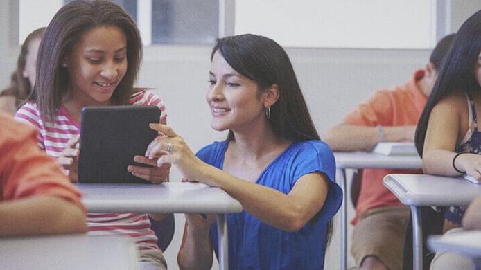 female pre k - 12 teacher helping a young female student on her tablet in classroom