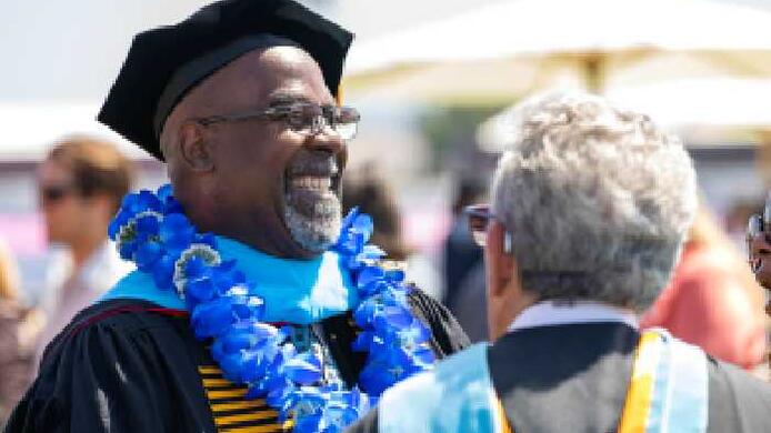 A man wearing graduation regalia associated with the Ed.D. degree smiles while standing next to another person.