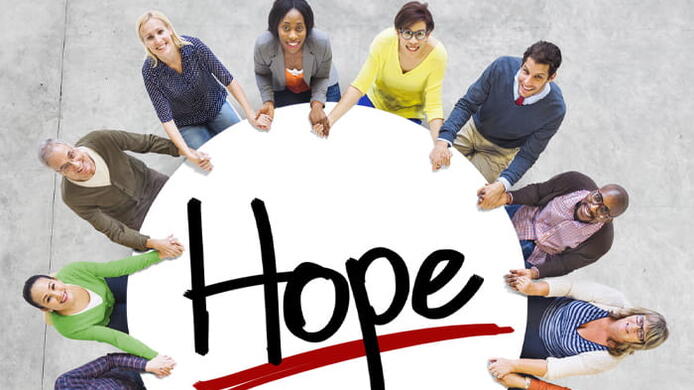 An overhead view of a diverse group of people encircling a round sign emblazoned with the word Hope, which is designed in a handwriting-style font.