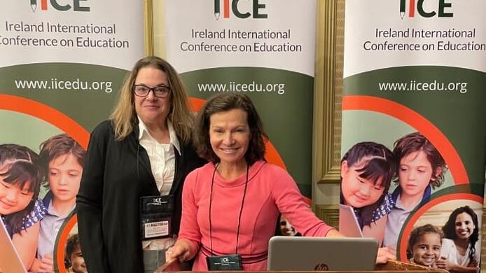 UMass Global faculty members Dr. Kimberly Greene and Dr. Jessica Bogunovich stand side by side behind a podium. Behind them are three identical posters that read IICE Ireland International Conference on Education and also contain the URL www.iicedu.org.