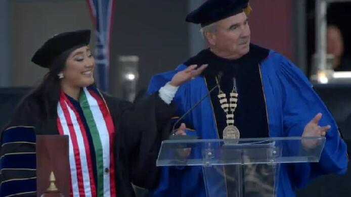 Grad and academic advisor Stephanie Herrera stands next to Chancellor David Andrews on the stage during the 2022 Southern California Commencement. She is pointing toward her parents, who are out of the picture. Both Herrera and Andrews wear graduation regalia.