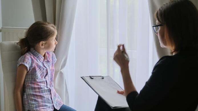 A girl looks at a window while interacting with a female social worker holding a clipboard.