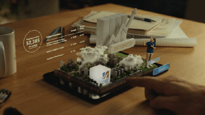 A screenshot from a UMass Global ad showing a small-scale human figure and imagery representing business education appearing as a 3D hologram from an electronic tablet.