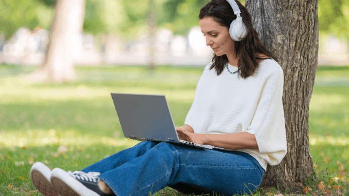 A woman wearing headphones using a laptop while sitting with her back to a tree trunk.