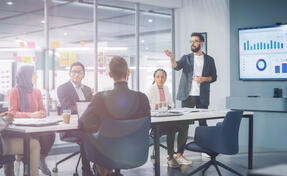 Diverse Modern Office: Businessman Leads Business Meeting with Managers, Talks, uses Presentation TV with Statistics, Infographics. Digital Entrepreneurs Work on e-Commerce Project.