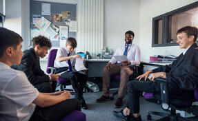 A small group of male secondary school students, sitting down in a circle and engaging in conversation with their teacher and each other in a meeting room. Some of them are looking at paperwork while they all discuss school topics.