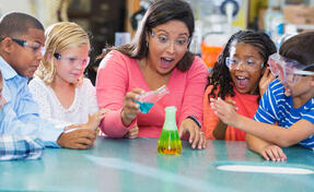 An Hispanic woman in her 40s teaching a multi-ethnic group of elementary school students in science lab. They are doing an chemistry experiment with colorful liquids in beakers.