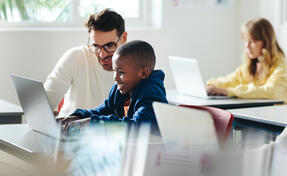 Male teacher helps a young boy with computer-based learning in a classroom setting. Child tutor providing a lesson in an elementary school, with a focus on coding and basic digital literacy.