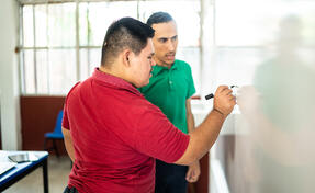 Young man with Down Syndrome is solving a math problem while his male teacher, who has a cognitive disability, looks on.