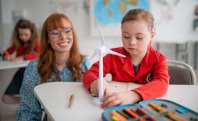 A down syndrome schoolgirl with model of wind turbine with help of teacher learning about eco-friendly renewable sources of energy in class at school, integration concept.