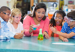 An Hispanic woman in her 40s teaching a multi-ethnic group of elementary school students in science lab. They are doing an chemistry experiment with colorful liquids in beakers.