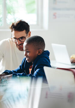Male teacher helps a young boy with computer-based learning in a classroom setting. Child tutor providing a lesson in an elementary school, with a focus on coding and basic digital literacy.