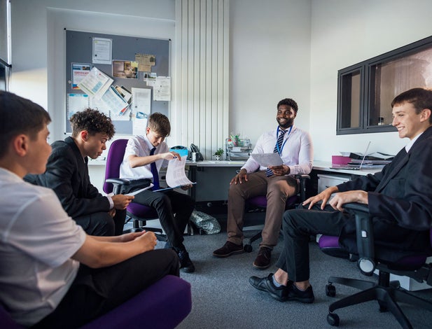 A small group of male secondary school students, sitting down in a circle and engaging in conversation with their teacher and each other in a meeting room. Some of them are looking at paperwork while they all discuss school topics.