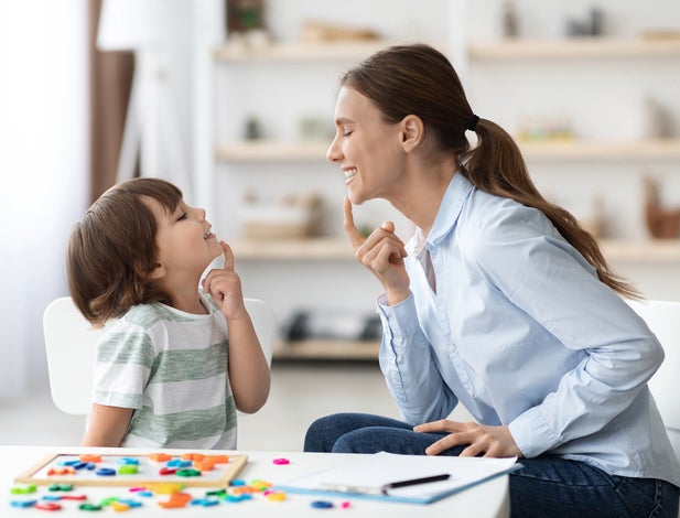 Speech training for kids. Professional woman specialist training with little boy at cabinet, teaching him right articulation exercises, side view