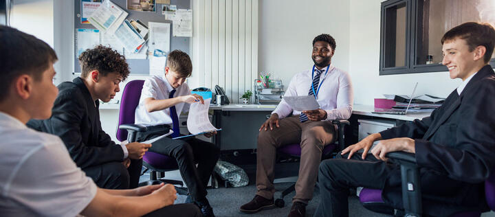 A small group of male secondary school students, sitting down in a circle and engaging in conversation with their teacher and each other in a meeting room. Some of them are looking at paperwork while they all discuss school topics.