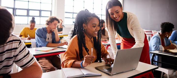 Smiling young female teacher helping a student working on a laptop during a lesson in a school classroom