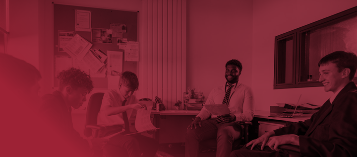 A small group of male secondary school students, sitting down in a circle and engaging in conversation with their teacher and each other in a meeting room. Some of them are looking at paperwork while they all discuss school topics.