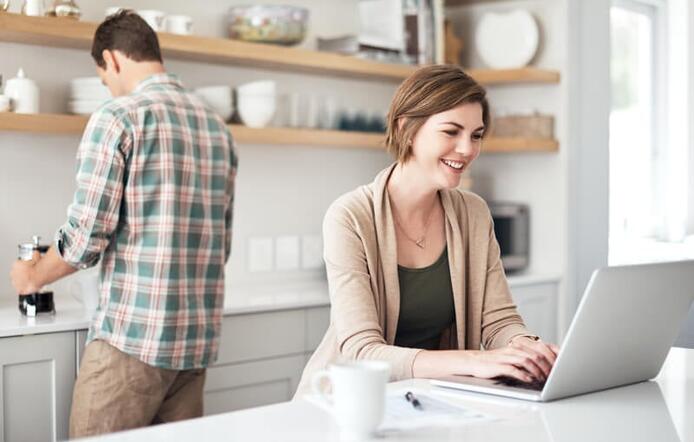 female at a laptop in the kitchen while male spouse prepares something behind her