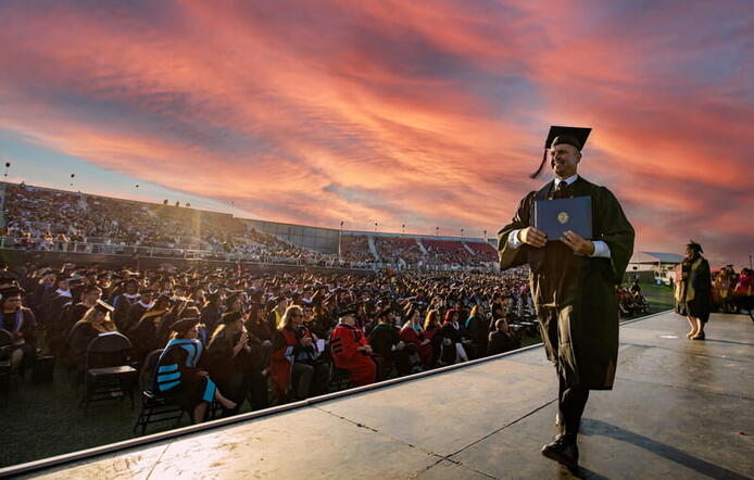 UMass Global Graduate walking across the stage with audience in the background