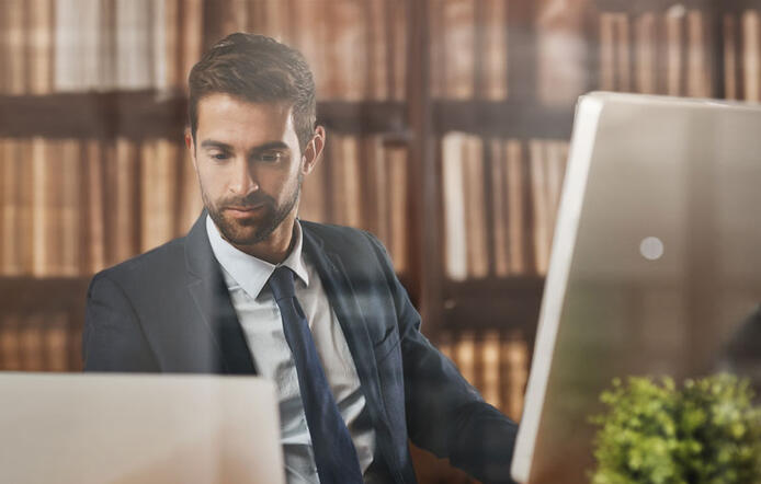 man working on a computer in a paralegal job with books behind him