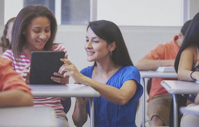 female pre k - 12 teacher helping a young female student on her tablet in classroom