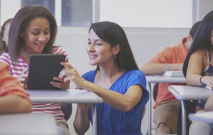 female pre k - 12 teacher helping a young female student on her tablet in classroom