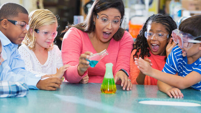An Hispanic woman in her 40s teaching a multi-ethnic group of elementary school students in science lab. They are doing an chemistry experiment with colorful liquids in beakers.